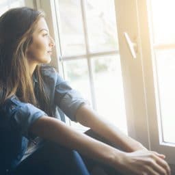 Smiling young woman sitting and looking through the window. With long hair, wears denim shirt. Hands on legs.