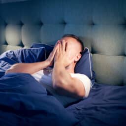 man holding head in hands, blue bedspread and a light illuminating his discomforted position
