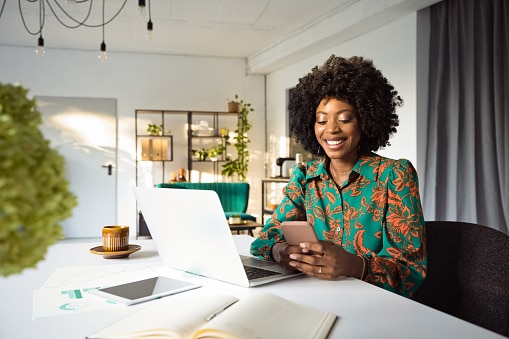Smiling tech-savvy woman working on a laptop and checking her phone