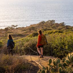 Two female friends hiking together at Torrey Pines State Park in San Diego California on a windy winter day, however it is still sunny and brights and you can see the Pacific ocean in the background